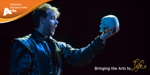 Male actor on a darkened stage facing toward the right with his left arm held out in front of him and looking at the skull he is holding in his left hand. Delaware Division of the Arts logo on an orange banner in the upper left hand corner and Bringing the Arts to Life is written in the lower right hand corner of the image.