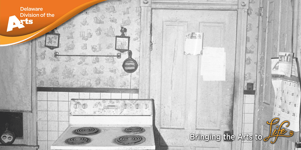 black and white photograph of an old unused kitchen with a vintage electric stove in front of a wall with tiles on the bottom half and wallpaper on the upper half next to a door