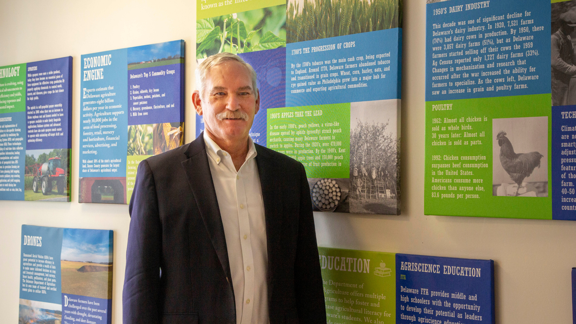 Delaware Secretary of Agriculture Michael T. Scuse stands in the agency lobby in front of colorful panels related to the history of Delaware agriculture.