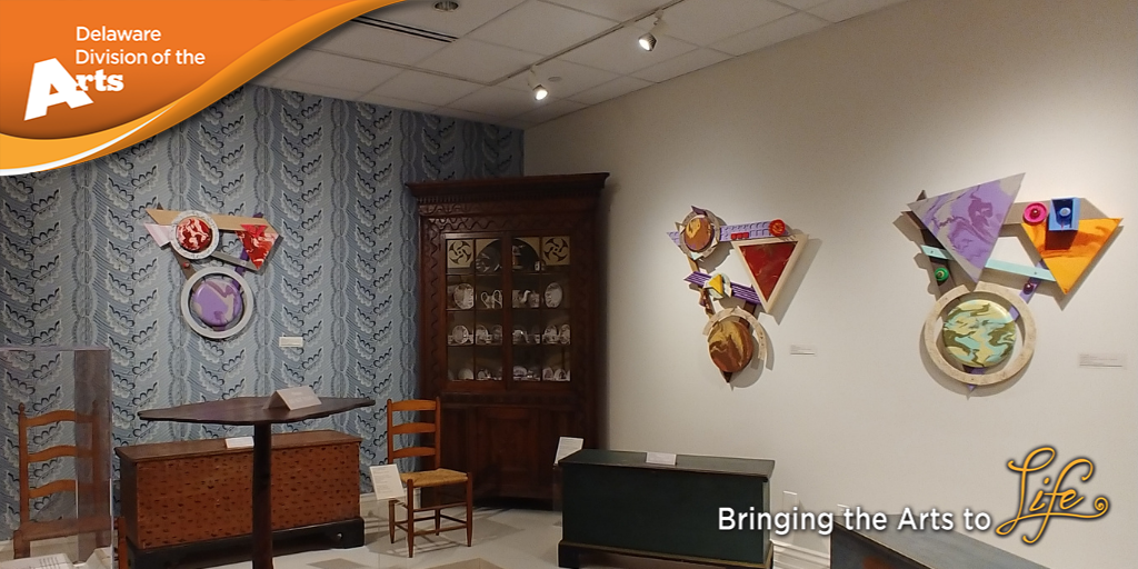 Delaware Division of the Arts orange logo banner over 2021 Individual Artist Fellow Jack Knight's artworks hanging on two walls among pieces of antique furniture on display in the Biggs Museum of American Art