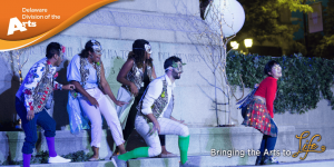 Delaware Division of the Arts orange logo banner and tagline of Bringing the Ars to Life over an image of Delaware Shakespeare's performance of a Midsummer Nights Dream performed outside in a park featuring four actors standing in a group wearing masks behind a female actor, all looking at the female actor and listening to her, while the female actor has her back turned towards the group of actors