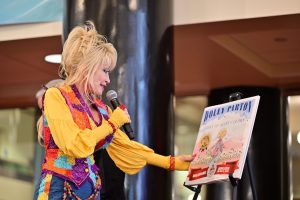 Dolly Parton stands holding a microphone and looking at a book positioned on an easel.