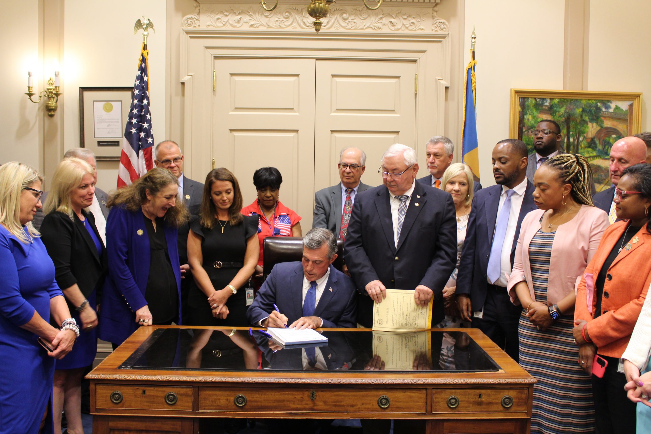 Governor Carney signs a bill at his desk while surrounded by members of the General assembly.