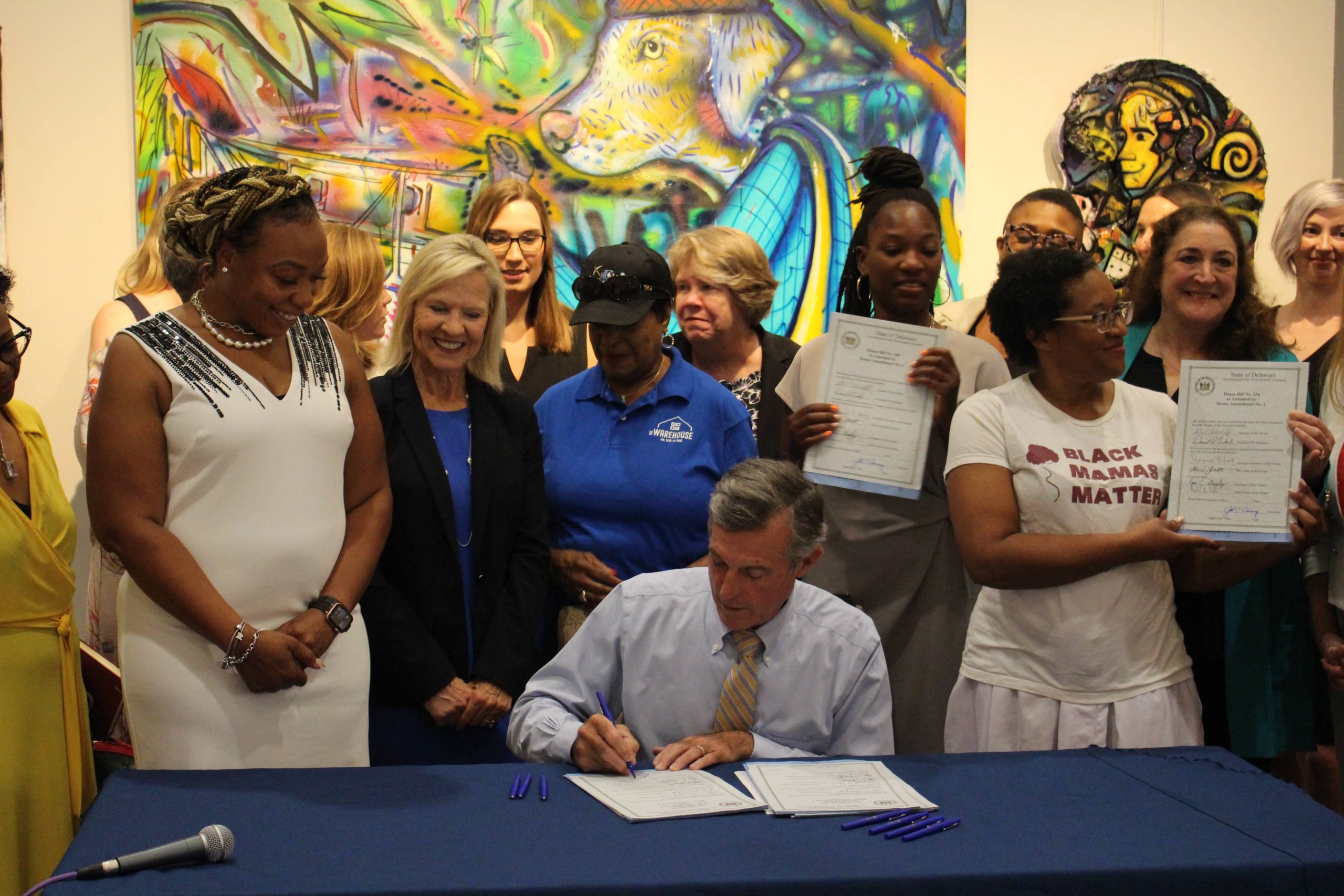 Governor Carney signs legislation while seated at a desk and supporters stand behind him.