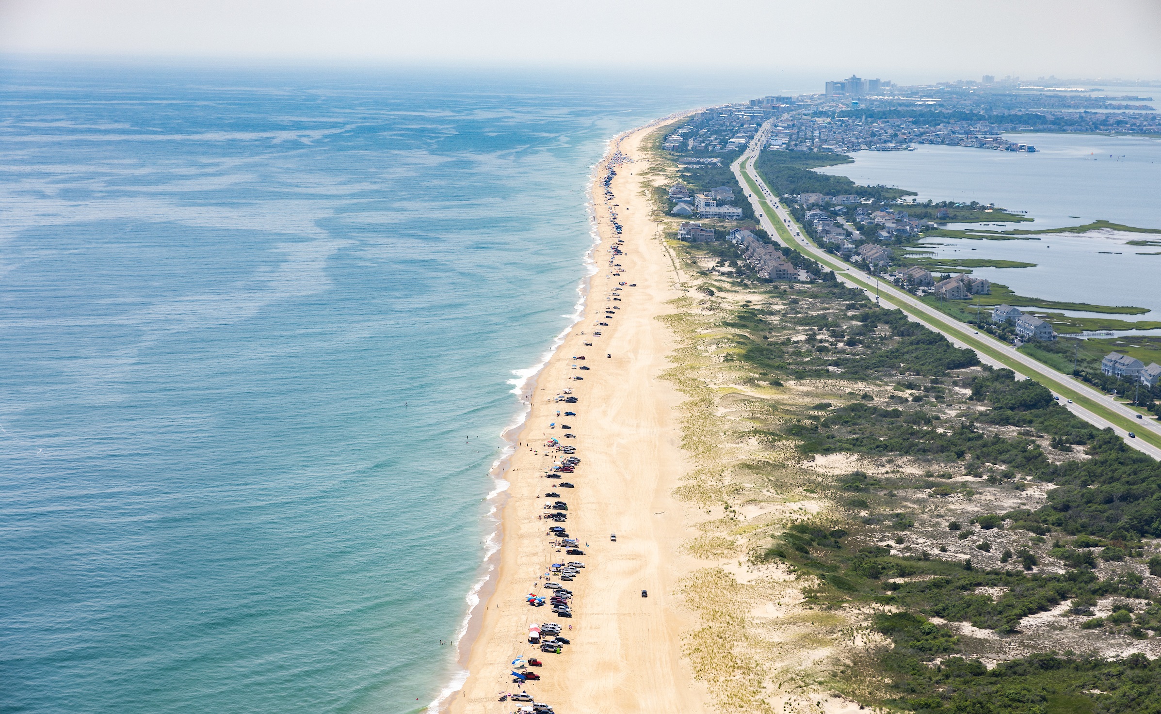 An overhead shot of the downstate Delaware coastline with drive-on surf anglers along the beach, which is pictured vertically up the center of the photo,with the Atlantic Ocean to the left and Route 1 surrounded by natural vegetation to the right.