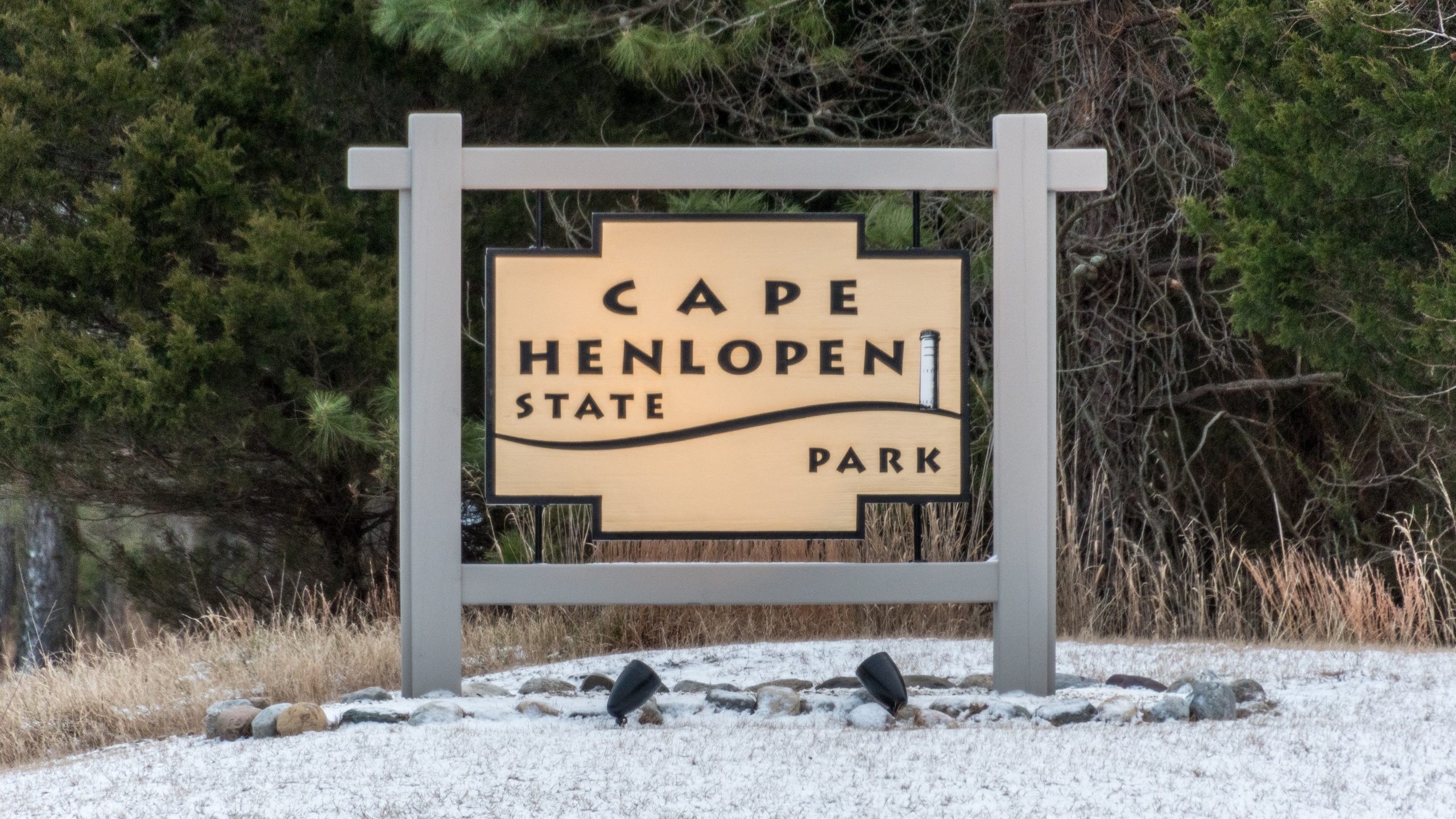 A wood Cape Henloepn State Park signs stands in the center of the photo surrounded by a snowy ground in the forefront and trees in the background.