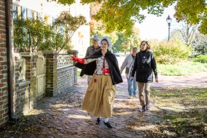 A woman dressed in a yellow 18th-century dress with a black cloak and red gloves leads a group of visitors to First State Heritage Park in Dover around the park's brick-lined sidewalks and historic buildings.