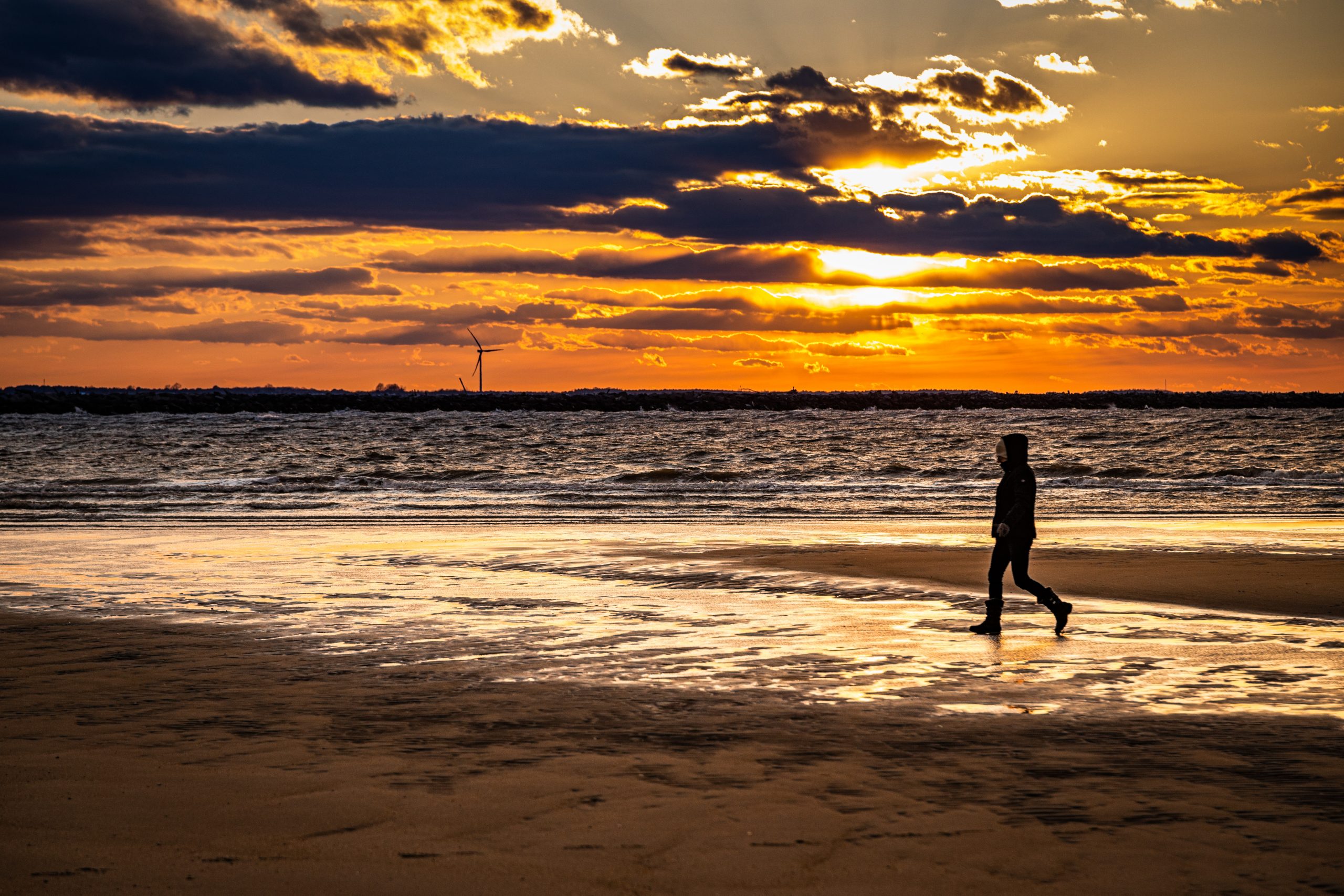 A photo of a person walking from the right side of the frame to the left on a beach with wet sand and a setting sun at The Point in Cape Henlopen State Park.