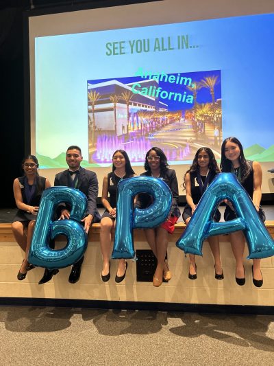 Six students sitting on the edge of a stage smiling while holding three large balloons that spell out BPA.