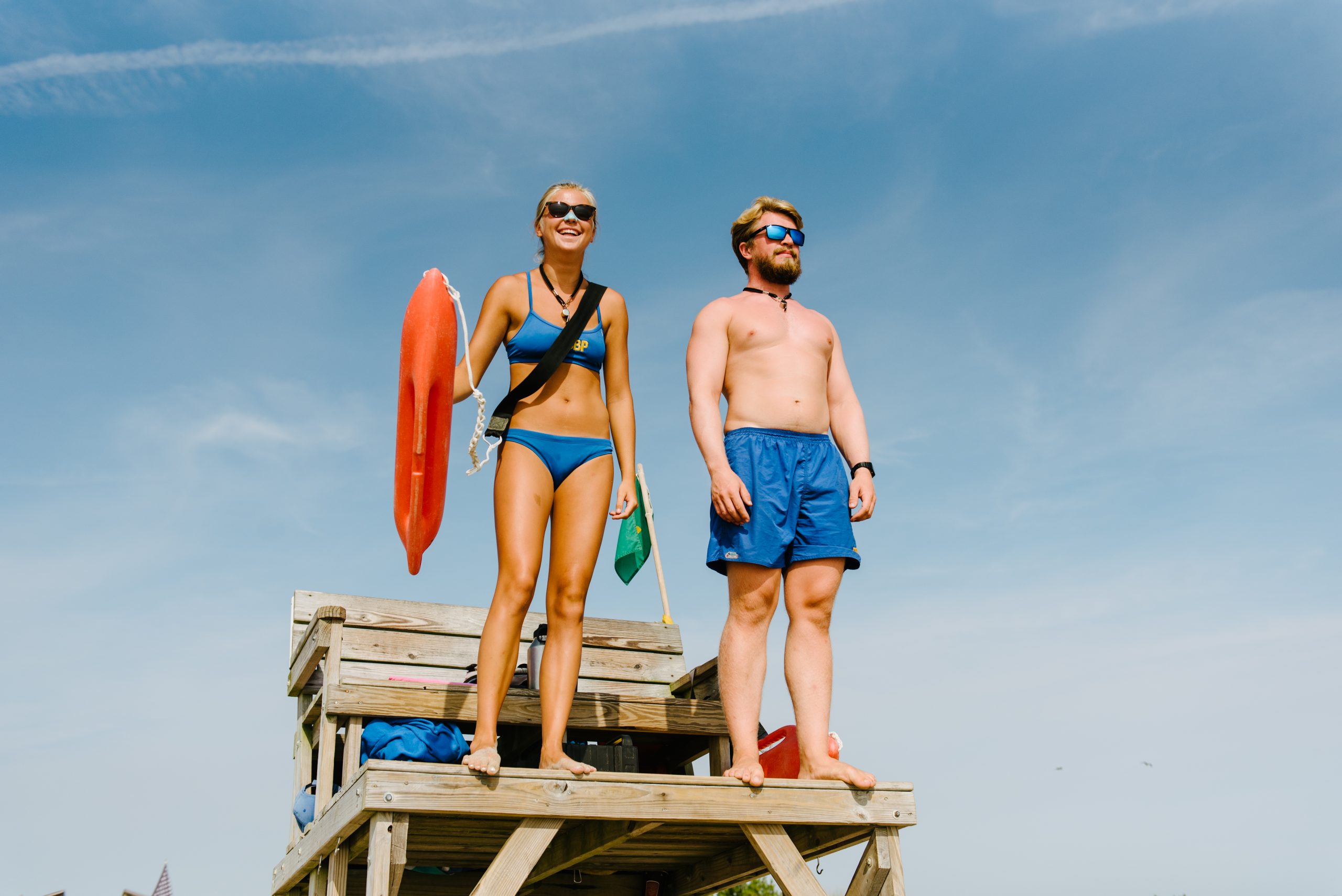 A team of lifeguards, one woman and one man, dressed in blue bathing suits stand on a lifeguard stand against a blue, slightly cloudy sky. The woman lifeguard is also holding a lifesaving floatation device.
