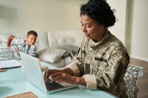 A woman in military fatigues is working at a computer in her home while a young child looks on.