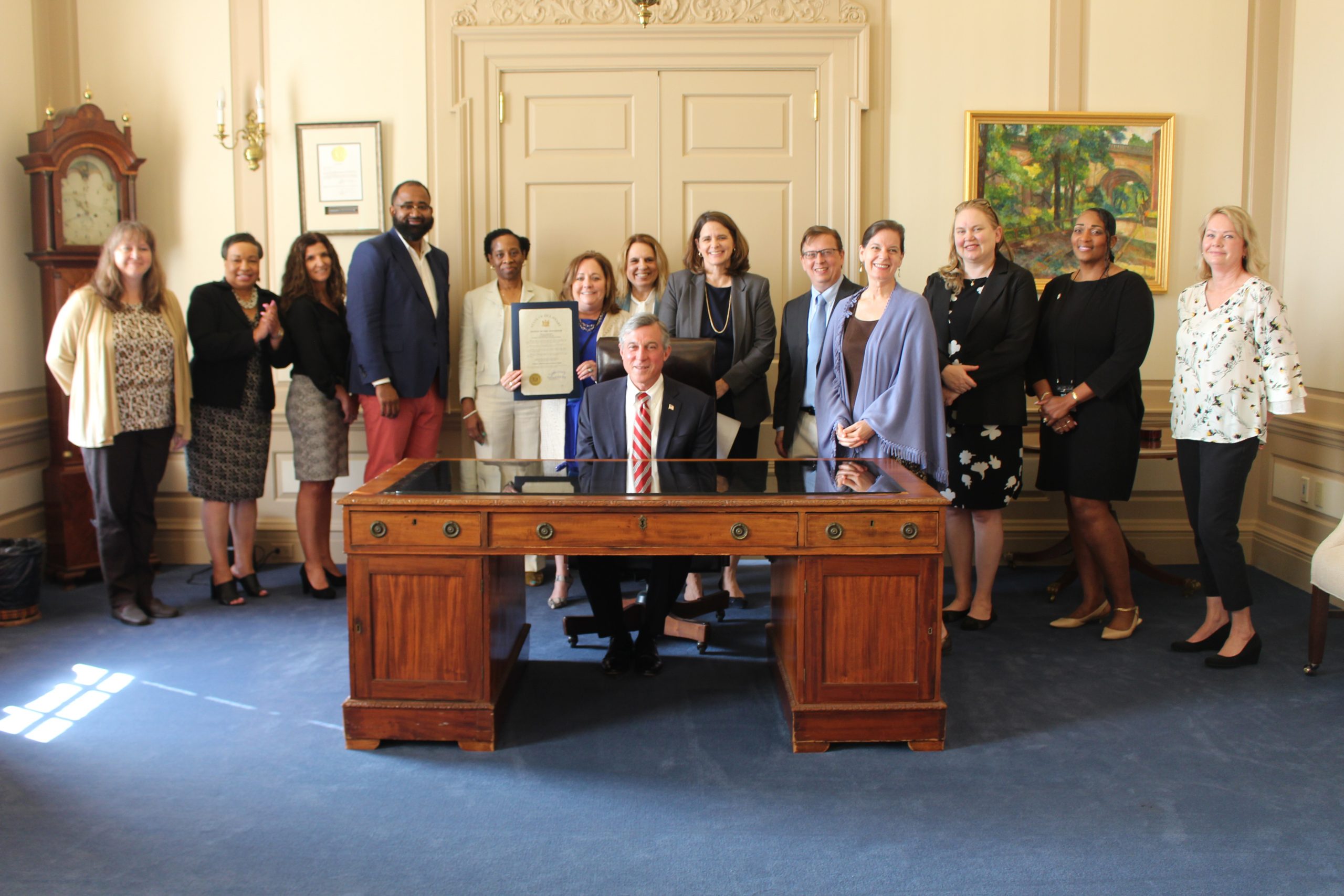 Governor Carney sits at his desk and is surrounded by employees of the Delaware Division of Human Resources. One of the employees holds up the proclamation for Public Service Recognition Week while everyone poses for a photo.