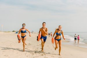 Three lifeguards, two females with a male in the center, in Delaware State Parks Beach Patrol blue uniform bathing suits run along the beach, each carrying an orange life saving devices.