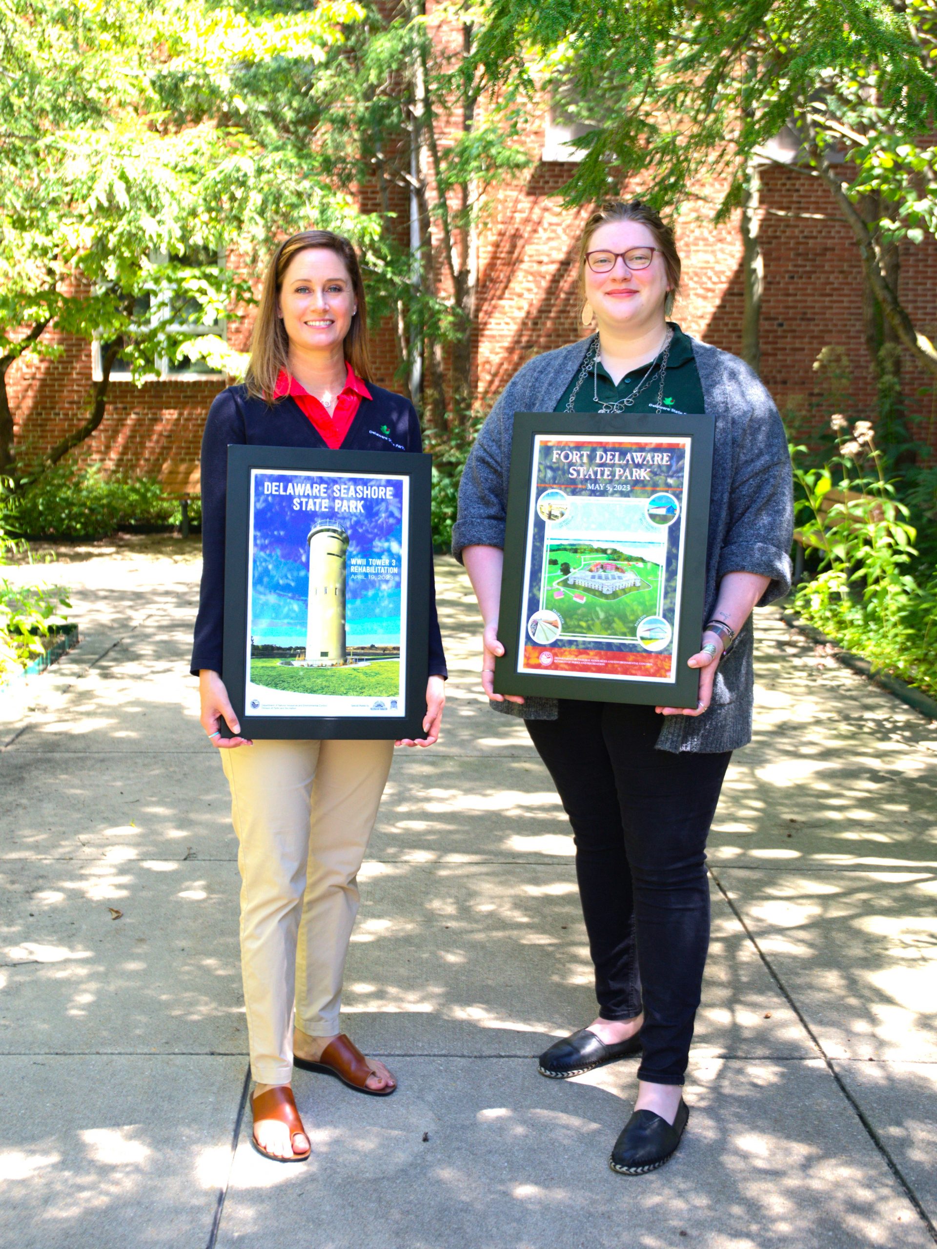 Two women stand on a sidewalk among trees holding framed posters, one of WWII Tower 3 at Delaware Seashore State Park and one of Fort Delaware State Park