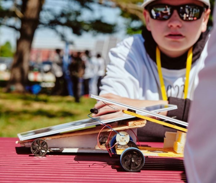 Boy with sunglasses on in front of a racer for the 2023 Junior Solar Sprint competition.
