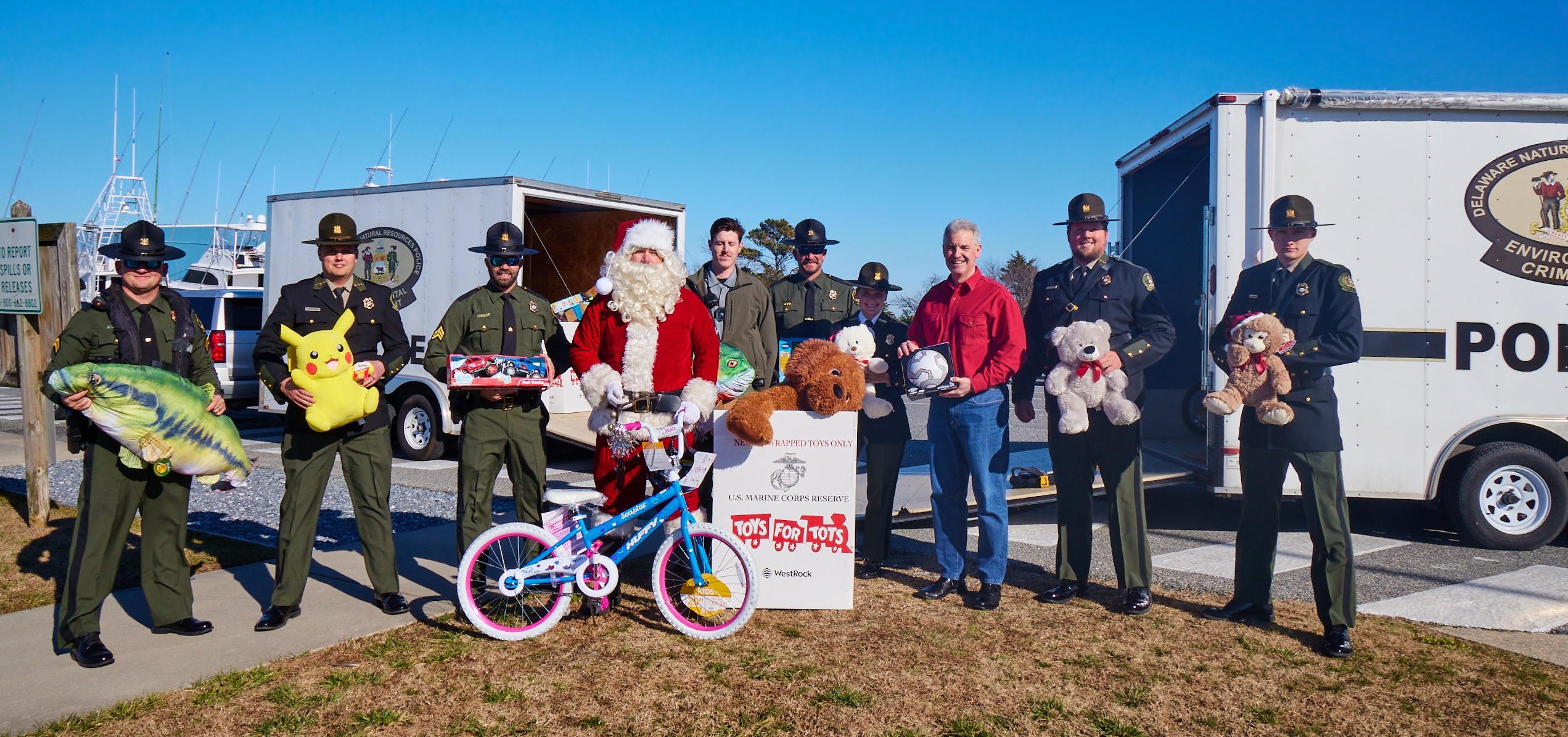 Delaware Natural Resources Police officers, DNREC Secretary Shawn M. Garvin and Santa stand holding toys behind a Toys for Tots donation box.