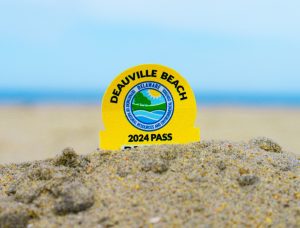 a yellow sticker with the words Deauville Beach 2024 Pass sticks out of the sand with a blurry blue sky and strip of blue ocean in the background.