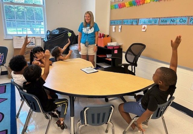 Students and staff sit around a table with small children raising their hands while a teacher watches.