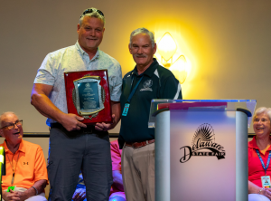 Dr. Mark Isaacs stands holding the Secretary's Award for Distinguished Service to Delaware Agriculture with Secretary Michael T. Scuse standing to his right, next to the Delaware State Fair podium.