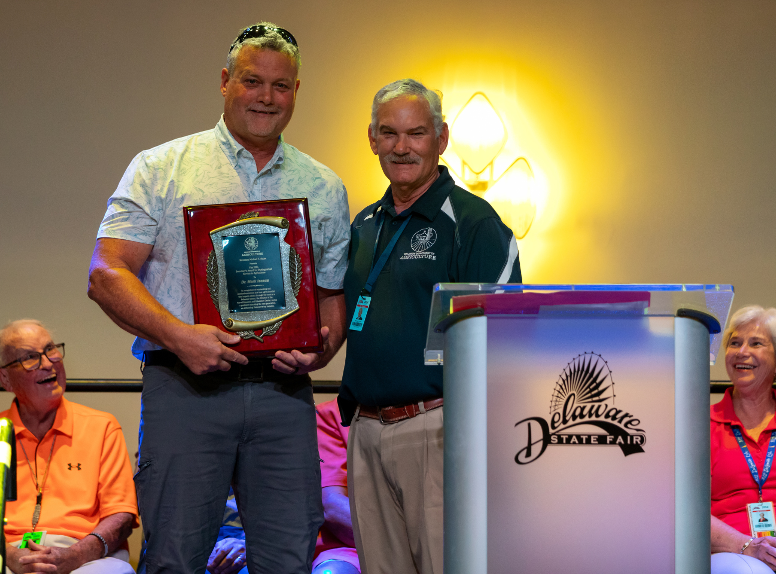 Dr. Mark Isaacs stands holding the Secretary's Award for Distinguished Service to Delaware Agriculture with Secretary Michael T. Scuse standing to his right, next to the Delaware State Fair podium.