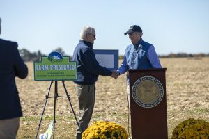 On a farm, Governor Carney shakes Secretary Scuse's hand after presenting him with the Order of the First State, with the new AgLands Farm Preserved sign in front.