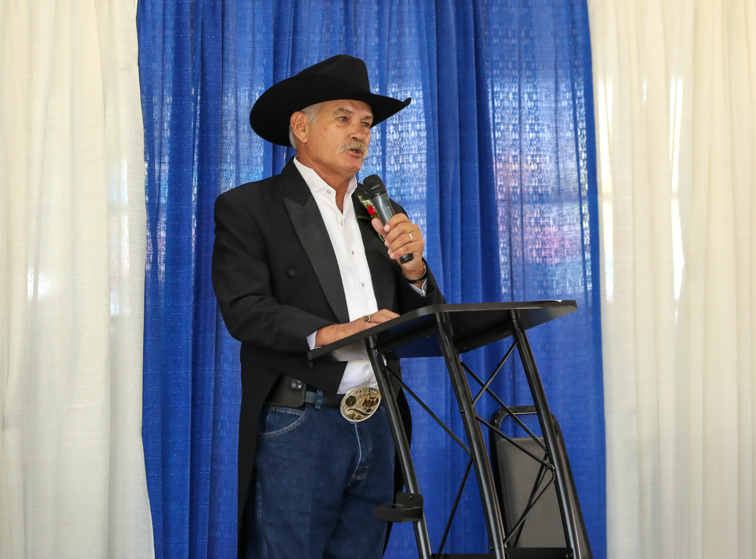 Secretary Scuse emceeing the Delaware State Fair Livestock Extravaganza in his black cowboy hat, tails, and blue jeans, with a blue and white pole and drape behind.