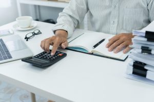 office employee works on desk, using calculator to manage financial documents and accounts, preparing an income tax report in a busy office setting.