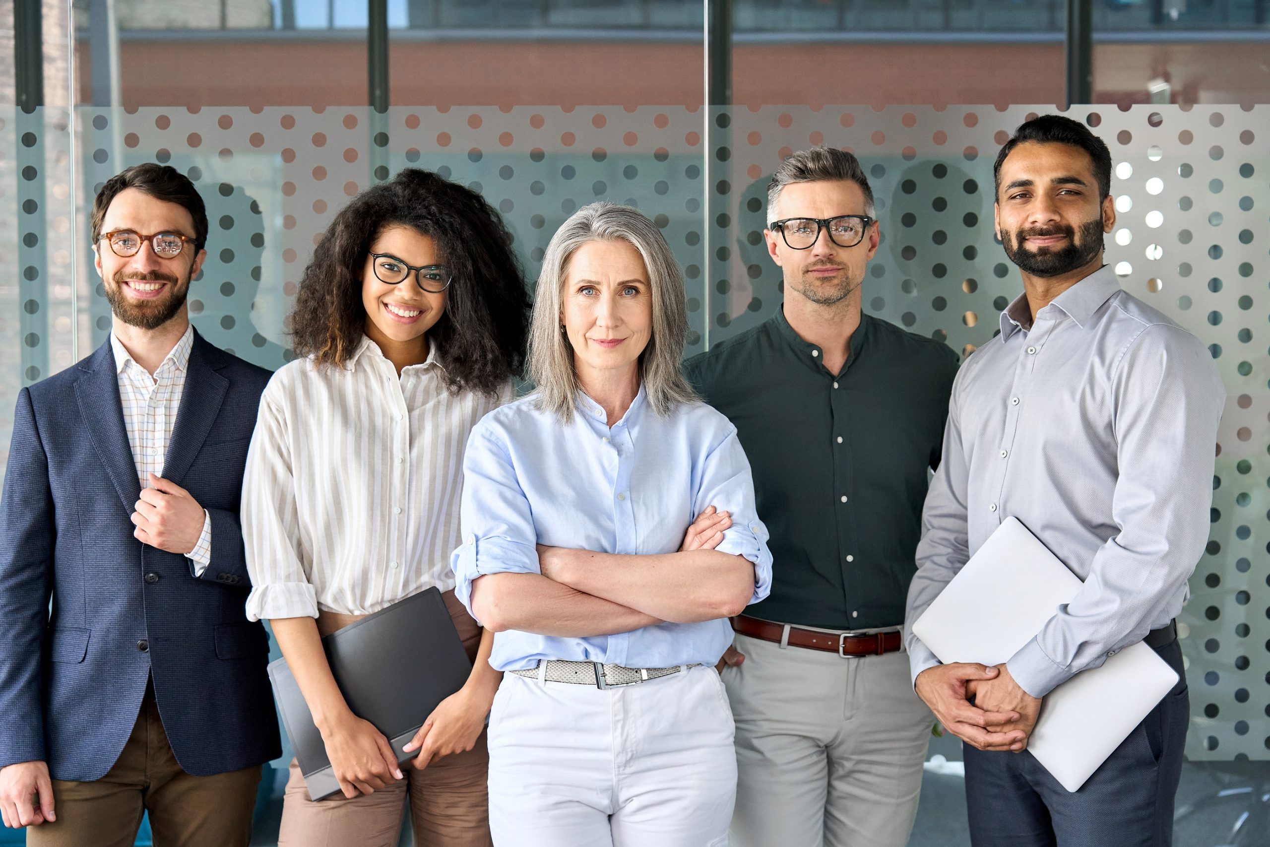 A diverse group of business professionals, including young multiethnic employees and an older executive leader, stand together in an office, smiling and looking at the camera.