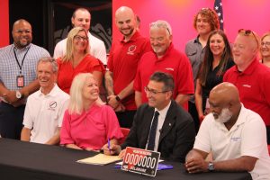 Governor Matt Meyer sits at a table smiling, signing a bill, with legislators, William Penn faculty and staff surrounding him.