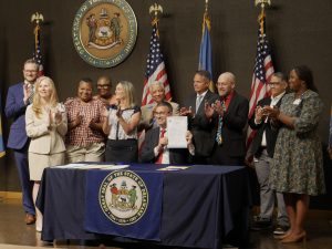 Governor Meyer sits behind a table with the State of Delaware seal in front, holding up a signed piece of legislation as legislators and advocates clap behind him.