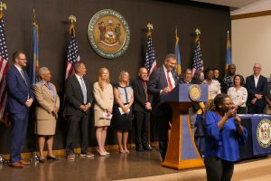 Governor Meyer stands behind a podium delivering remarks with legislators and advocates standing behind him.