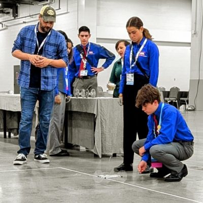 One student squats near the ground, pushing a robotic car, while five students and a teacher look on.