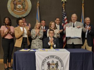Governor Matt Meyer sits at a table clapping with other legislators and advocates as Rep. Frank Burns holds up his signed bills to the right of the Governor.