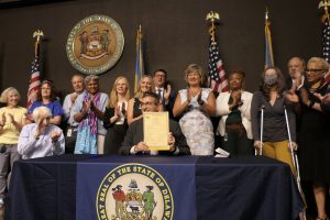 Governor Meyer sits at a table holding Senate Bill 4 with legislators and advocates standing behind him clapping.