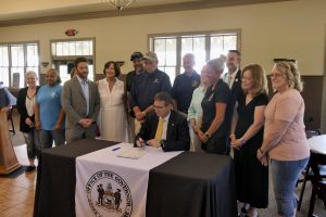 Governor Meyer sits at a table with the Governor's seal as he signs House Bill 187 with lawmakers and advocates standing behind him.
