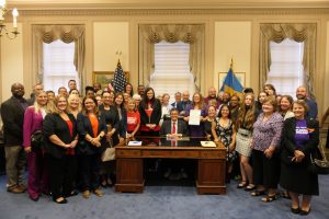 Governor Matt Meyer sits at his desk surrounded by dozens of lawmakers and advocates following the signing of HB 54.