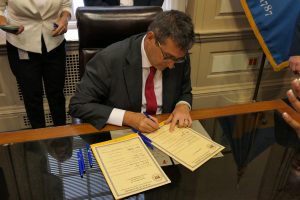 Governor Matt Meyer sits at his desk signing one of two bills laid in front of him.