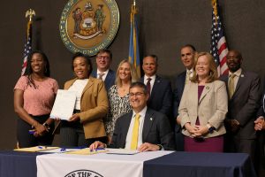 Governor Meyer sits at a table after signing a piece of legislation, surrounded by legislators and the bill sponsor Speaker Melissa Minor-Brown holding the bill up to the left of the Governor.