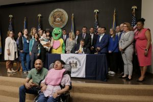 Governor Meyer sits at a table holding a signed bill with dozens of legislators and advocates standing behind him in applause.