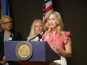 First Lady Lauren Meyer stands at a podium with Sen. Nicole Poore and DHR Secretary Yvonne Gordon behind her.