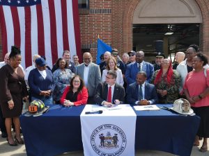 Governor Matt Meyer sits at a table signing legislation with legislators, other elected officials, advocates, and volunteer firefighters behind him.