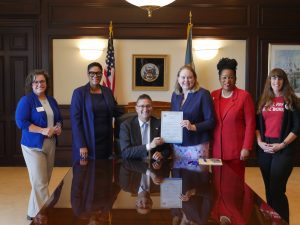Governor Matt Meyer sits at a conference table holding House Bill 105 with Rep. Melanie Ross Levin and other advocates.