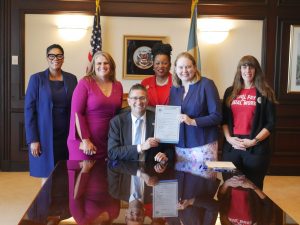 Governor Matt Meyer sits at a conference table holding House Bill 136 with Rep. Melanie Ross Levin and other advocates.