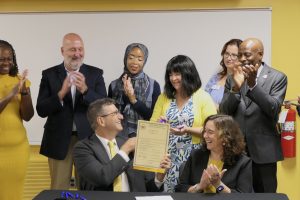 Governor Matt Meyer sits at a table holding a piece of signed legislation, handing it back to the sponsor Rep. Kim Williams as Secretary of Education Secretary Cindy Marten sits next to him clapping, and other legislators and advocates stand behind them celebrating.