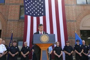 Governor Matt Meyer stands at a podium with a large vertical American flag hanging behind him and a line of volunteer firefighters.