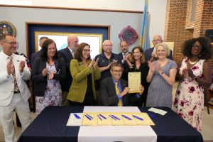Governor Meyer holds up a piece of legislation that he just signed as legislators, state leaders, and advocates stand behind him, clapping. 