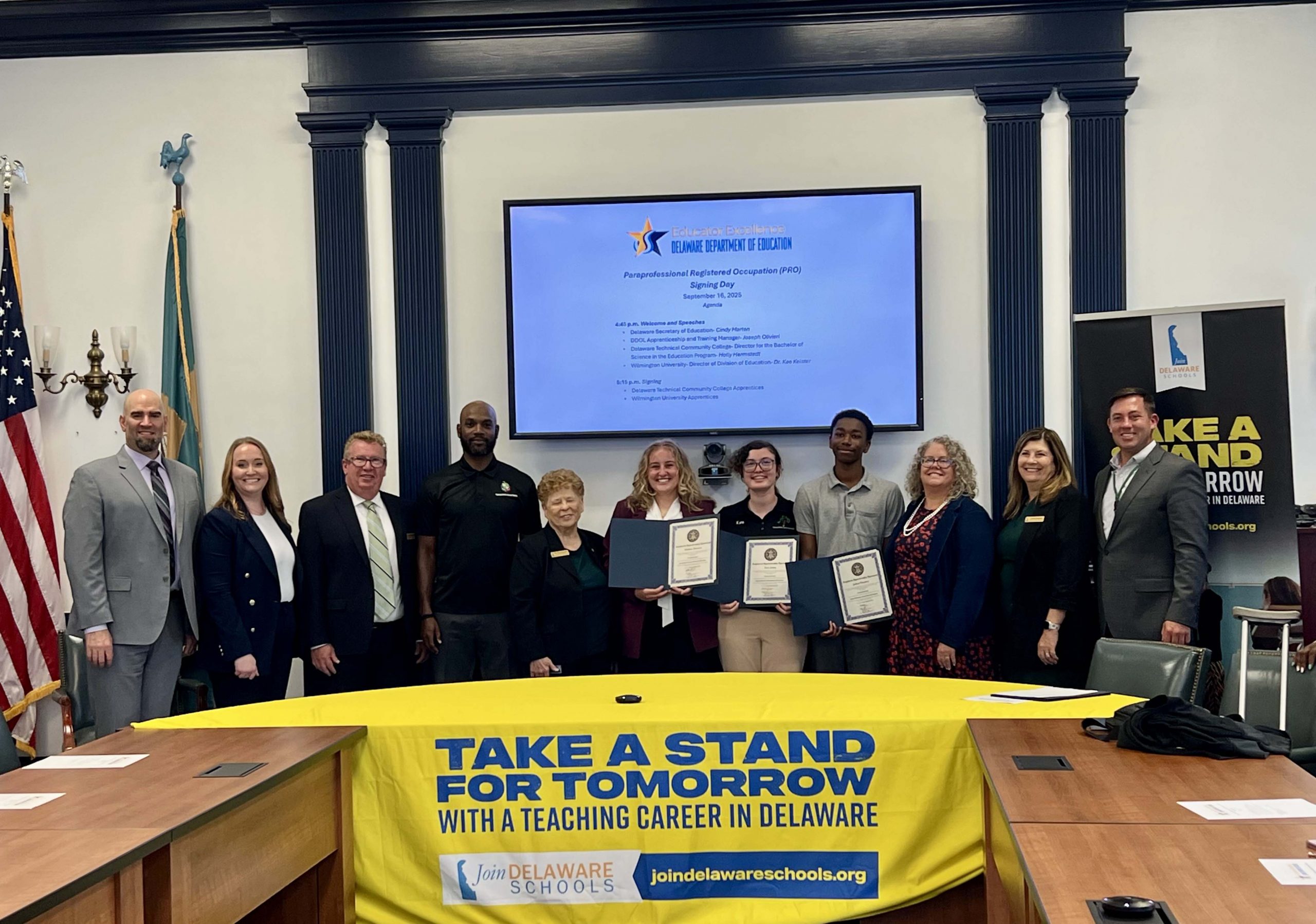 Smiling Department of Education staff and others in a large conference room with a man and two women holding certificates.