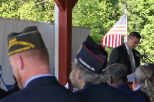 Governor Matt Meyer stands in front of a podium with an American flag in the background. Servicemembers sit in the foreground. 