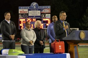 Governor Matt Meyer stands at a podium on a football field. Legislators and advocates stand behind him. You can see the scoreboard in the background. 