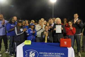 Governor Matt Meyer sits at a table on a football field after signing a ceremonial copy of a bill. Dozens of advocates and legislators stand behind him clapping. An advocate to the right of Governor Meyer's holds the bill. 